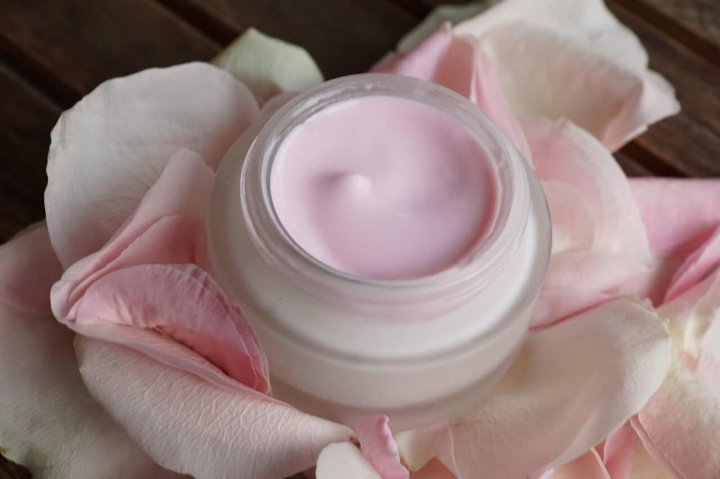 Pink rose face cream in a glass jar surrounded by fresh rose petals.